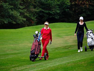 Two women walking on a golf course in sports attire, pulling golf carts. The grass is lush and green, with trees lining the background under a clear sky.