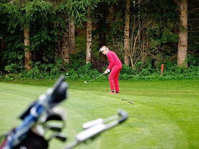 A golfer in a red outfit prepares to swing on a green course with trees lining the background and a golf bag in the foreground.
