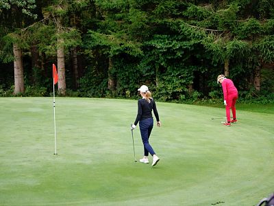 Two people on a golf course green, one preparing to putt and another standing by. Trees in the background create a peaceful setting.