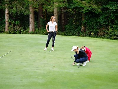 Three women on a golf course green; two kneeling and focusing on putting, one stands observing. Background features lush trees and clear sky.