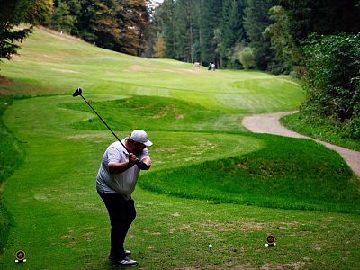 A golfer swings his club on a green golf course, surrounded by trees. He is wearing a cap, and the surroundings are lush and serene.