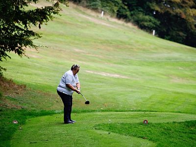 Woman on golf course preparing to tee off, surrounded by lush green landscape and trees. Casual attire, holding a club, ready to swing.