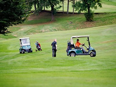 Green golf course with two golf carts and trees in the background. A person is walking near a golf cart, likely preparing to play.