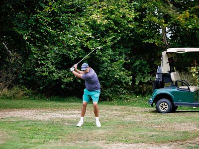 A man in a blue cap and green shorts is swinging a golf club on a grassy course with a golf cart nearby, surrounded by lush trees.