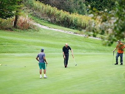 Three men are playing golf on a grassy course surrounded by trees. One is putting the ball, while the others watch.