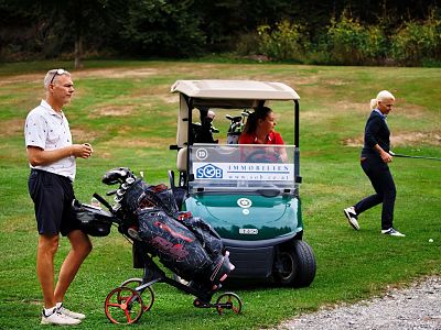 Three people at a golf course: a man near a golf cart, a woman inside the cart, and another woman preparing to swing a golf club.