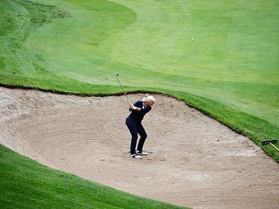 Golfer in a sand trap on a lush golf course swings his club, with green grass and a rake nearby.