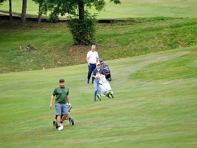 Four people on a golf course, pulling carts over green grass, surrounded by trees and a gentle slope, under a clear sky.