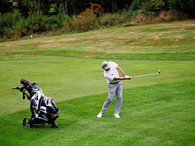 A golfer in a white shirt and gray pants swings a club on a green golf course, with a golf bag and another person watching in the background.