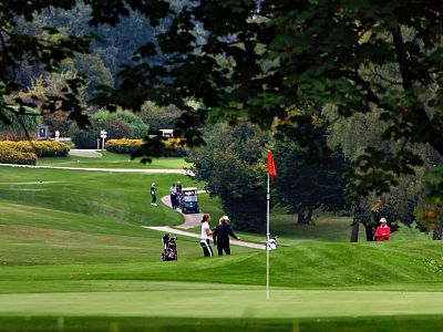 A peaceful golf course scene with green fairways, trees, golfers, and a red flag marking the hole, under a clear sky.