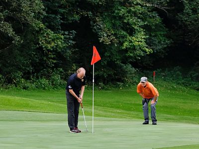 Two golfers on a green course, one in a black shirt putting, the other in orange watching, surrounded by trees. The green is lush and the flag is red.
