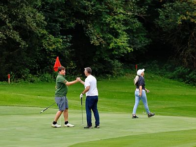 Two golfers shake hands on a green golf course, while another person walks nearby. Lush greenery and trees surround the scene.
