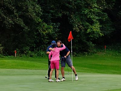 Three golfers in colorful outfits hug near a flag on a golf green, surrounded by lush trees and grass, celebrating a successful game.
