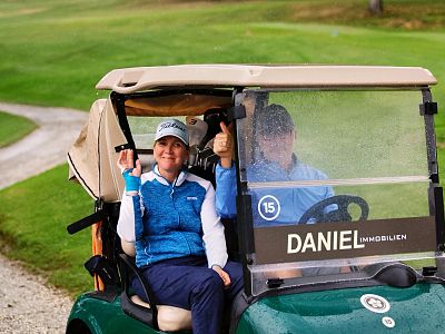 Two golfers in a cart on a lush course, one giving a thumbs-up, with 'DANIEL IMMOBILIEN' sign on cart, cloudy sky in the background.