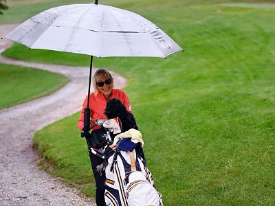 A golfer with a large umbrella walks on a rainy golf course path, pushing a golf cart with a bag, surrounded by green grass.