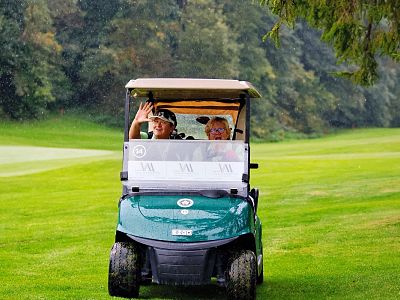 Two people wave from a green golf cart on a lush, green golf course with trees in the background, creating a relaxed outdoor setting.