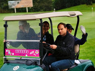 A group of three people sitting in a green golf cart with a pink advertisement. One person waves while raindrops cover the cart's windshield on a golf course.