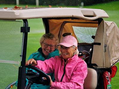 Two women in colorful raincoats smile while sitting in a golf cart on a rainy day at a golf course.