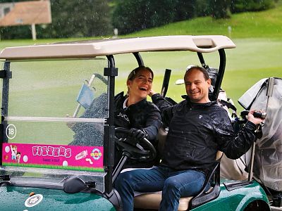 Two people in raincoats enjoying a laugh in a green golf cart during rain on a golf course.