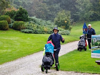 Two golfers in rain gear walk on a path at a green golf course, pushing trolleys with clubs, as rain falls around them.