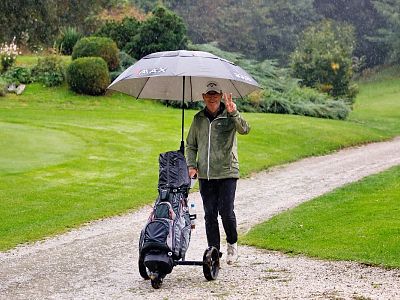 Person walking on a golf course path with an umbrella in the rain, carrying golf equipment. The surrounding area is green and lush.