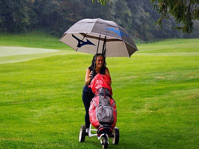 Woman on a rainy golf course pushing a cart under an umbrella, smiling next to a green field and a distant flag.