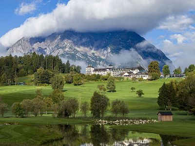 Lush green fields with a reflective pond, quaint building, and a cloud-covered mountain in the background under a blue sky.