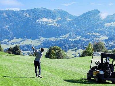 A golfer swings on a green fairway with mountains in the background. A golf cart is nearby. The sky is clear and blue, providing a serene atmosphere.