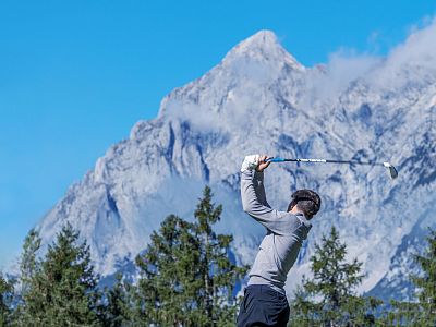 A golfer mid-swing on a course with towering mountains and cloudy blue skies in the background.