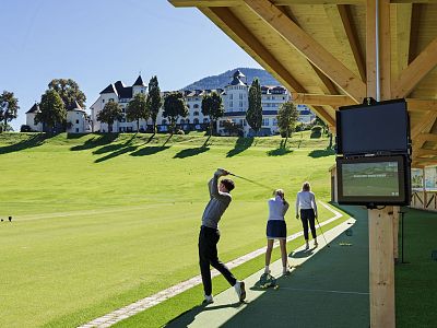 Three golfers at a driving range surrounded by greenery; a large, white building and mountains are visible in the background.