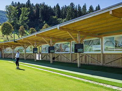 A covered golf driving range with wooden structures and green grass in a picturesque setting, surrounded by trees and hills under a blue sky.