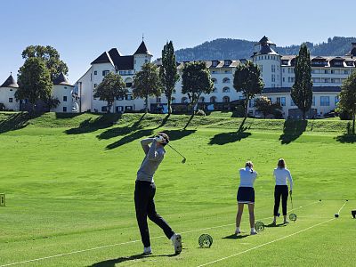 Golfers on a lush green course with a large traditional building and hills in the background, practicing their swings under clear skies.