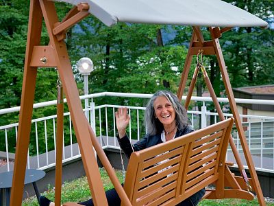 Woman with gray hair sitting on a wooden swing on a terrace, smiling and waving, surrounded by lush green trees and white railing.