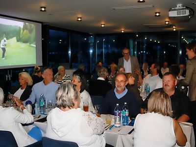 People seated at a formal dinner event indoors. A projector displays a golfer on a screen. Attendees are engaged in conversation around tables.