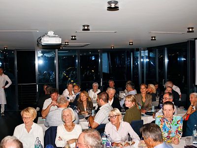 People seated at tables in a dimly lit room, engaged in conversation, at a formal event. Attendees dressed in smart casual and formal attire.