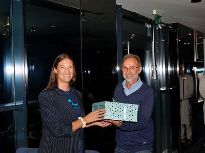 Two people exchanging gift boxes at an event. They are smiling, suggesting a friendly and warm atmosphere. The setting appears to be modern and professional.