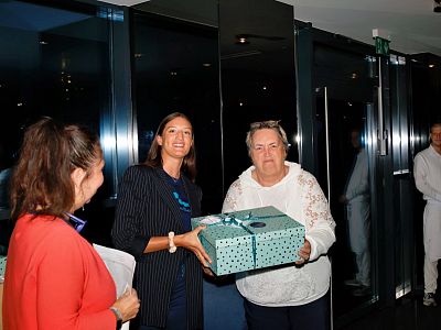 Two women in formal wear present a gift box to another woman indoors. Reflections are visible on the glass wall in the background.