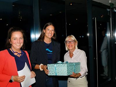 Three women pose together indoors, smiling. One holds a gift box with a ribbon. They appear relaxed and are in a modern room with glass walls.