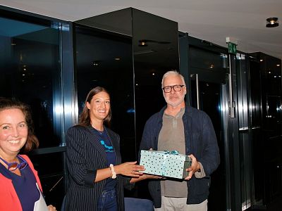 Three people in a dimly lit room, two women and a man. One woman presents a gift box to the man. They all smile, standing near a reflective wall.