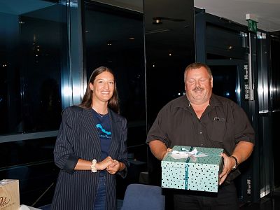 Two people in a dimly lit room, one holding a gift box wrapped in blue with a ribbon. They are smiling and standing side by side. The background is dark.