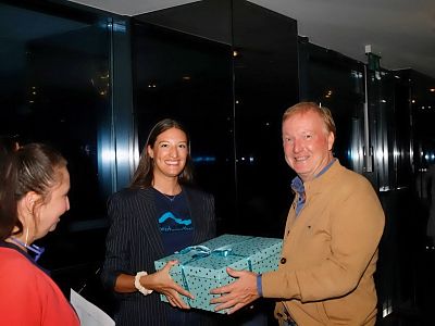 Three people share a joyful moment exchanging a gift, with one person handing over a teal gift box to another. They are in a dimly-lit, elegant setting.