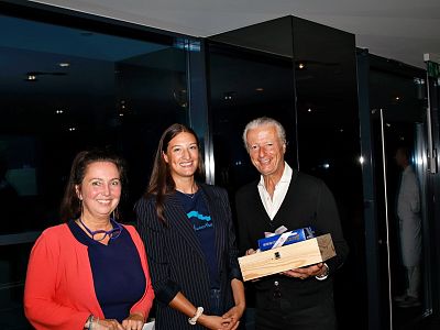 Three people at an indoor event, smiling. Two women stand next to a man holding a wooden box, all dressed in business-casual outfits.