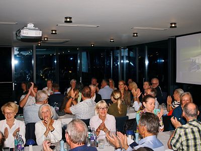 Group of people in a room clapping and smiling, seated around tables during an event. The dim lighting and projector suggest a presentation or award ceremony.