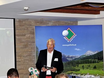 Man in suit presents at a golf event with a scenic mountain landscape and golf logo in the background.