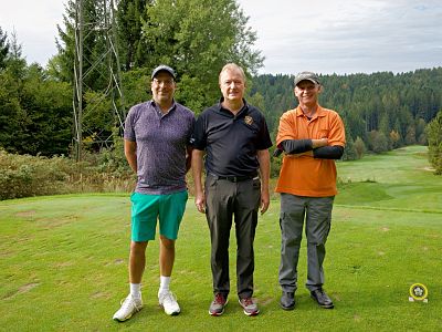 Three men stand on a golf course, smiling, with trees and green fairway behind them. They wear casual golf attire, including shorts and caps.