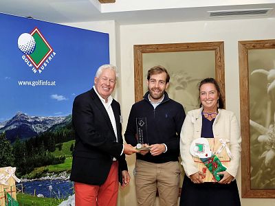 Three people at an indoor event with a golf theme, smiling and holding awards and gifts, with a scenic banner in the background.