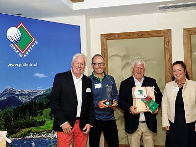Four people at a golf award ceremony, two holding trophies, with a golf course and mountain backdrop. Everyone is smiling.