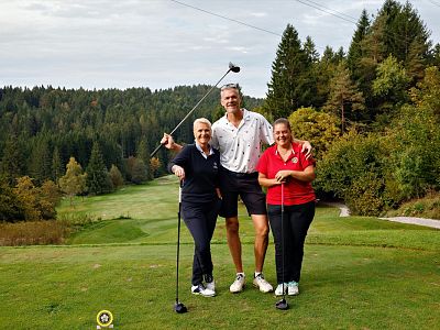 Three people stand on a golf course with trees in the background, holding golf clubs and smiling at the camera.