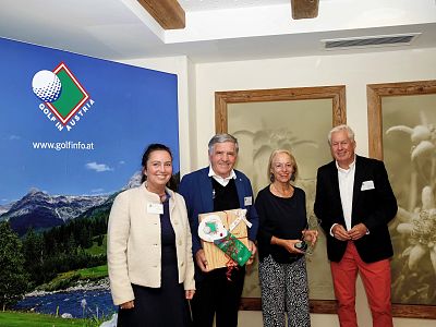 Four people at an event, two holding a gift and certificate, with a golf-themed backdrop and framed pictures on the wall.