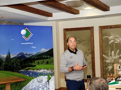 A man speaks in a room with wooden accents, standing in front of a screen displaying a golf-related image. There's a person seated in the foreground.